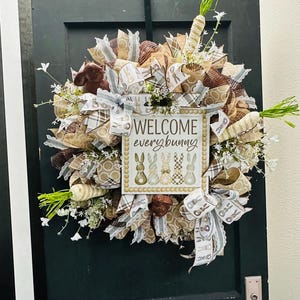 Peut inclure: Une couronne de P&acirc;ques avec une pancarte &laquo; Welcome every bunny &raquo;. La couronne est compos&eacute;e de rubans bruns, beiges et blancs, de carottes d&eacute;coratives et de petites fleurs blanches. Accroch&eacute;e &agrave; une porte vert fonc&eacute;.