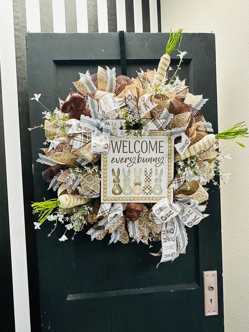 Peut inclure: Une couronne de P&acirc;ques avec une pancarte &laquo; Welcome every bunny &raquo;. La couronne est compos&eacute;e de rubans bruns, beiges et blancs, de carottes d&eacute;coratives et de petites fleurs blanches. Accroch&eacute;e &agrave; une porte vert fonc&eacute;.