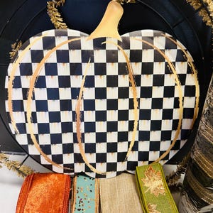 May include: A flat pumpkin-shaped sign featuring a black and white checkered pattern with gold-colored brushstroke outlines. Below the sign are four rolls of ribbon in orange, teal with gold glitter, tan, and green with gold leaf patterns.