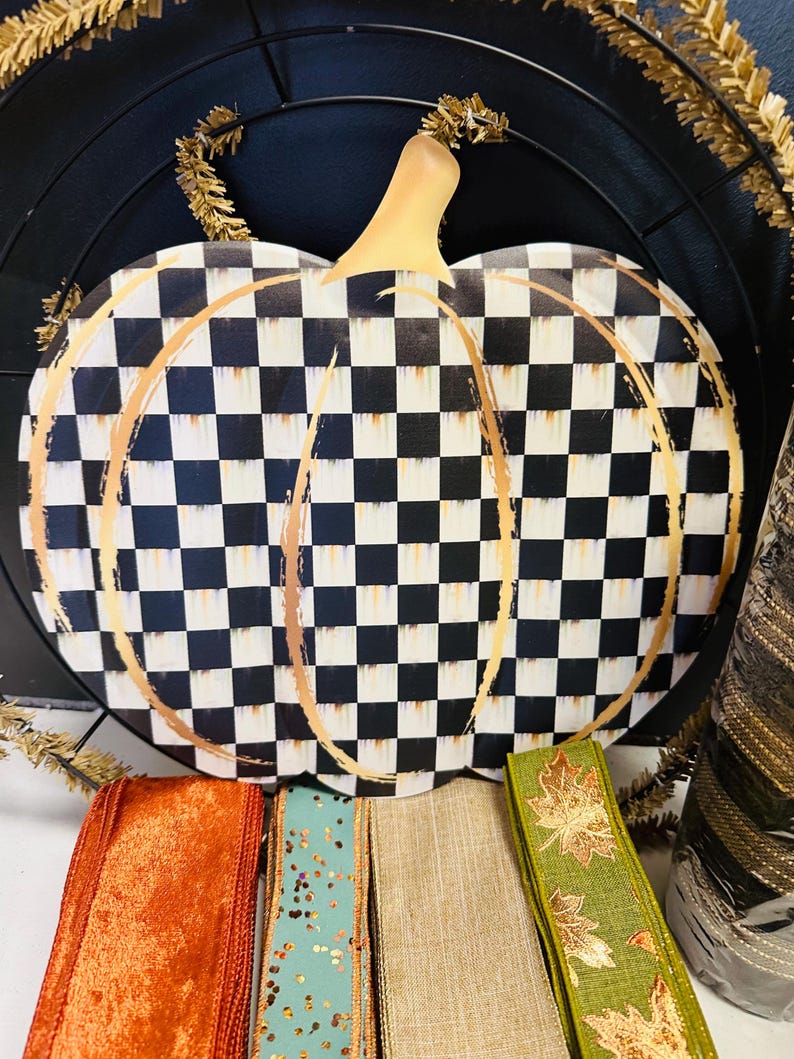 May include: A flat pumpkin-shaped sign featuring a black and white checkered pattern with gold-colored brushstroke outlines. Below the sign are four rolls of ribbon in orange, teal with gold glitter, tan, and green with gold leaf patterns.