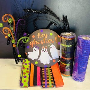 May include: A Halloween-themed wreath with a black base, adorned with orange and green decorative elements. A round sign with the text "Hey ghosties!" and illustrations of ghosts is featured. Rolls of black and purple mesh and colorful ribbons complete the festive arrangement.