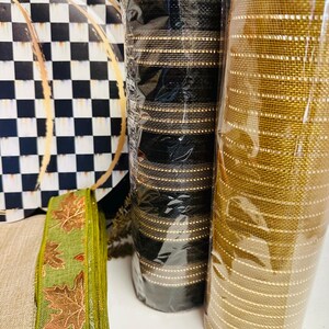 May include: Two rolls of mesh ribbon, one black with gold stripes and one solid gold, stand next to a green ribbon featuring a copper-colored leaf pattern and a round sign with a black and white checkered pattern and gold line accents.