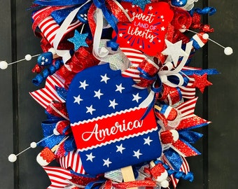 May include: A red, white, and blue patriotic door wreath with a popsicle-shaped sign that says "America" and a red sign that says "Sweet Land of Liberty".