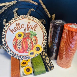 以下が含まれることがあります： ヒョウ柄の縁取りと「hello fall」の文字が入った丸い看板が、カボチャとヒマワリを囲んでいます。秋色のメッシュロールや様々なリボンも見えるので、秋をテーマにしたクラフトプロジェクトのようです。