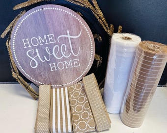 May include: A round wooden sign with the text HOME SWEET HOME, a black wire wreath frame with gold accents, four rolls of patterned fabric ribbon in tan and white, and two rolls of mesh material in white and tan with a grid pattern.