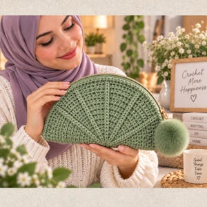 Op de afbeelding: Een saliegroene halfronde gehaakte etui met een gestructureerd waaiervormig steekpatroon, een goudkleurige ritssluiting en een bijpassende pluizige pompon aan de zijkant. Het accessoire heeft een gestructureerd, afgerond silhouet.