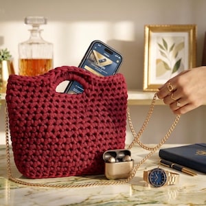 Könnte beinhalten: Eine weinrote Häkeltasche mit goldener Kette, in der sich ein Smartphone befindet. Neben der Tasche befinden sich eine goldene Uhr, kabellose Ohrhörer und ein blaues Notizbuch. Eine Hand mit Ringen hält die Kette. Der Hintergrund umfasst eine Karaffe und gerahmte Kunst.