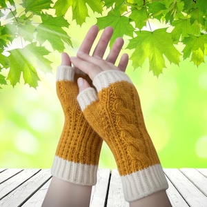 May include: Mustard yellow fingerless gloves with a cable knit design and off-white cuffs. The gloves are made of a soft, knitted material. The background is a blurred green and white.
