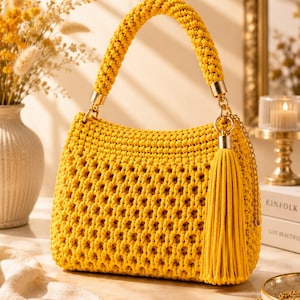 Può includere: Una borsa a mano in macramè giallo brillante caratterizzata da un motivo a trama aperta e testurizzata, un manico spesso intrecciato e una lunga nappa abbinata fissata a un anello in metallo dorato. La borsa ha una forma rettangolare.