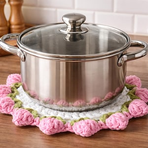 May include: A stainless steel pot with a glass lid sits on a handmade crochet trivet. The trivet features a white center with a border of pink and green crocheted flowers. The pot has two handles and a shiny finish.