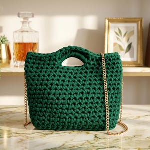 Può includere: Borsa tote verde scuro, lavorata all'uncinetto a mano, con tracolla a catena dorata. La borsa ha una forma rettangolare con un manico nella parte superiore. La borsa è appoggiata su una superficie di marmo.
