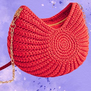 May include: A vibrant red crocheted handbag with a gold-colored zipper and chain strap. The bag features a circular design on the front and a curved shape. The chain strap is attached to the bag with gold-colored hardware. The background is a light purple with white and blue paint splatters.