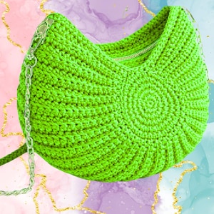 Op de afbeelding: Felgroene gehaakte schoudertas met een zilveren ketting en ritssluiting. De tas heeft een spiraalvormig ontwerp aan de voorkant en is gemaakt van getextureerd garen. De achtergrond heeft een aquareleffect met gouden accenten.