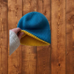 Könnte beinhalten: Ein Wendemütze in Blau und Gelb. Die Mütze ist aus weichem Strickmaterial gefertigt. Die blaue Außenseite kontrastiert mit der gelben Innenseite. Die Mütze wird vor einem Holzgrund gezeigt.