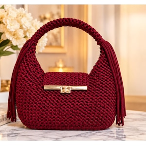 May include: A deep burgundy crochet handbag featuring a rounded silhouette, a single top handle, and long fringe accents on both sides. The bag includes a rectangular gold-tone metal clasp closure centered on the front flap, showcasing a textured knit.