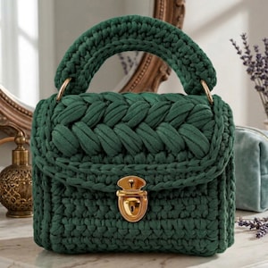 May include: A dark green crochet handbag featuring a thick braided top handle and a textured knit body. The bag is secured with a polished gold-tone metal push-lock clasp attached to the front flap, providing a structured and compact appearance.