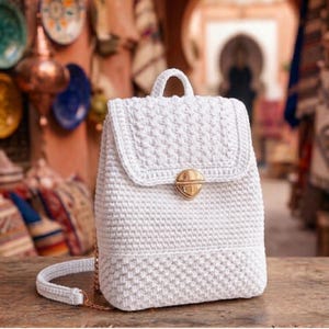 May include: A white crocheted backpack featuring a textured stitch pattern, a top handle, and a gold-toned metal push-lock closure on the front flap. The bag has a structured rectangular shape with a matching crocheted shoulder strap attached.