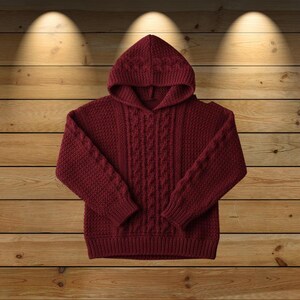 Könnte beinhalten: Burgunderfarbener Kapuzenpullover mit Zopfmuster auf der Vorderseite und den Ärmeln. Der Pullover ist aus einem dicken, strukturierten Strickstoff gefertigt und hat einen gerippten Saum und Bündchen. Die Kapuze ist ebenfalls gestrickt.