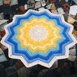 May include: A handmade, star-shaped crochet rug in shades of cream, yellow, and blue. The rug features a concentric design with a cream-colored center, transitioning to yellow, then blue, and a white border. The rug is placed on a patterned surface.
