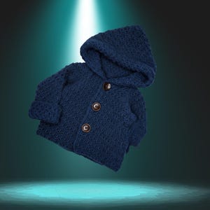 Könnte beinhalten: Eine marineblaue Kapuzenjacke aus Häkelarbeit für ein Kind. Die Jacke hat einen strukturierten Stich, lange Ärmel und drei braune Knöpfe auf der Vorderseite. Die Kapuze ist befestigt und die Jacke scheint handgefertigt zu sein.