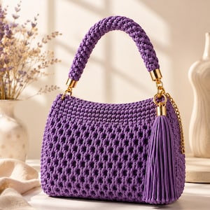 Può includere: Una borsa a mano in macramè viola con un motivo intrecciato testurizzato, un manico spesso intrecciato e una lunga nappa abbinata fissata a un anello in metallo dorato. La borsa ha una forma rettangolare strutturata con dettagli in metallo.