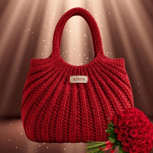 May include: A red, hand-knitted tote bag with a ribbed design and two handles. The bag has a rectangular tag that reads "AZVOS". A bouquet of red roses is visible in the lower right corner. The bag is made of a thick, textured yarn.
