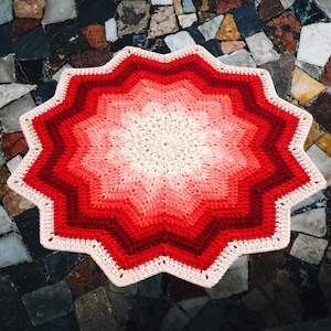 May include: A handmade, star-shaped crochet rug with a gradient of red hues, transitioning from a cream center to deep red edges. The rug features a scalloped border and is likely made of soft yarn.