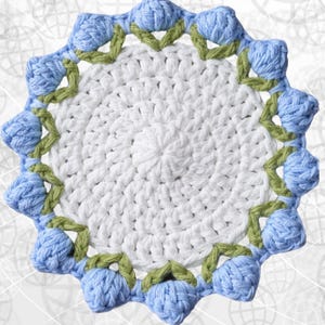 May include: A handmade, round crochet doily. The center is white, surrounded by a border of light blue flower-shaped accents and green leaves. The doily is a decorative item, perfect for adding a touch of handmade charm to any space.