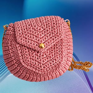May include: A dusty rose crochet handbag with a gold chain strap and a gold button closure. The round bag has a flap closure and a textured, woven appearance. The chain is a bright gold color.