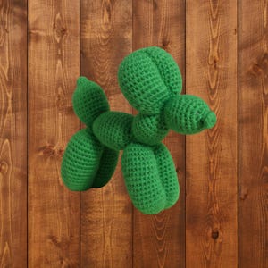 May include: A handmade green crochet dog, resembling a balloon animal, is displayed against a wooden background. The dog has a rounded head, elongated body, and four legs. The crochet stitches are visible, giving it a textured appearance. The dog is a whimsical decorative item.