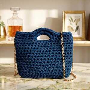 May include: A navy blue crochet handbag with a gold chain strap. The bag has a rectangular shape with a handle at the top. The background includes a shelf with a decanter, framed art, and books.