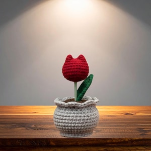 Puede incluir: Un tulipán rojo tejido a ganchillo con un tallo y una hoja verdes, plantado en una maceta gris. La maceta tiene un borde festoneado. La flor se exhibe sobre una superficie de madera. La maceta mide unos 10 cm de altura.