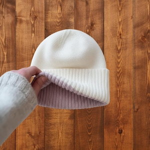 Peut inclure: Un bonnet réversible blanc et lavande. Le chapeau est fait d'un tricot côtelé doux. Le bord est replié, révélant l'intérieur lavande. Le chapeau est présenté sur un fond en bois.