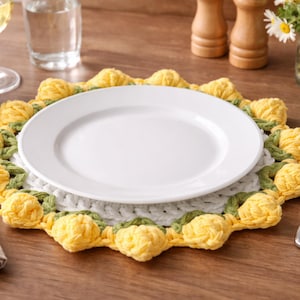 May include: A white ceramic plate rests on a handmade crochet placemat. The placemat features a white center, green leaves, and yellow flower-like accents. The placemat is on a wooden table, with a glass of water and other table settings in the background.