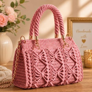 May include: A dusty pink crochet handbag featuring a textured geometric pattern on the front, two sturdy braided top handles with gold-tone hardware, and a detachable gold-tone chain shoulder strap. The bag has a structured rectangular shape.