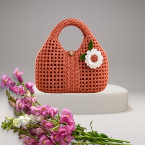 Può includere: Una borsa all'uncinetto color corallo, di forma arrotondata e con un unico manico. La borsa presenta un fiore decorativo bianco e marrone con una foglia verde. Un rametto di fiori rosa e bianchi è appoggiato davanti alla borsa.