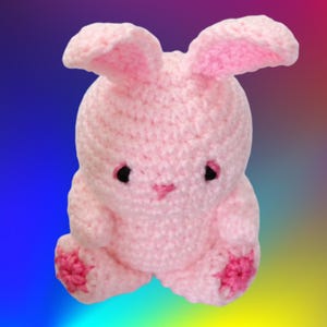 May include: A handmade, light pink crochet bunny with floppy ears, black eyes, and a pink nose. The bunny's paws are also detailed with pink accents. This soft toy is a charming example of handcrafted amigurumi.
