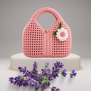 May include: A pink crocheted handbag with a floral embellishment and a decorative button closure. The bag has a structured shape with a handle and a lattice design. A sprig of lavender is in the foreground.