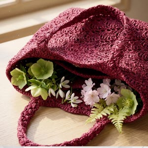 May include: A burgundy woven tote bag filled with an assortment of fresh flowers. The bag has braided handles and is open, revealing green, white, and pink blossoms. The bag is sitting on a light-colored wooden surface.
