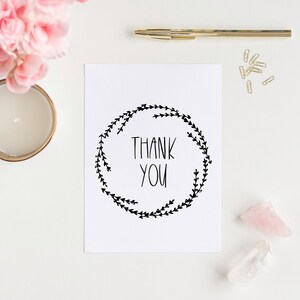 Puede incluir: Una tarjeta blanca con un diseño de corona negra y el texto "THANK YOU" en el centro. La tarjeta está sobre una superficie blanca con flores rosas, una vela dorada, clips de papel dorados y cristales rosas y transparentes.