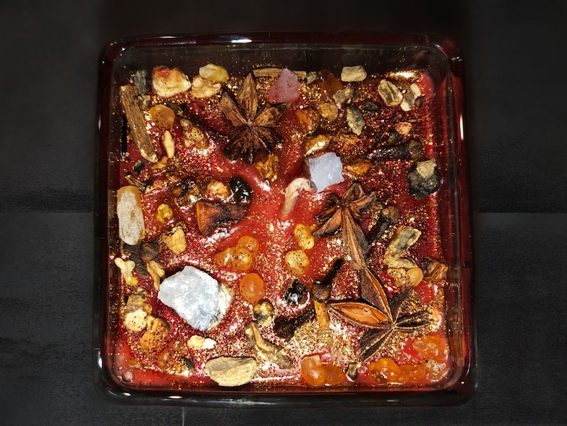 Peut inclure: Une bougie rouge avec des paillettes dor&eacute;es et divers &eacute;l&eacute;ments naturels incrust&eacute;s, notamment de l'anis &eacute;toil&eacute;, des b&acirc;tons de cannelle et des morceaux d'ambre. La bougie est dans un r&eacute;cipient en verre carr&eacute;.