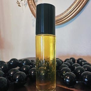 Puede incluir: Un frasco roll-on de vidrio transparente con tapa negra, lleno de líquido dorado. La botella está centrada, con una cadena de cuentas negras en primer plano. Un espejo con marco dorado está en el fondo.