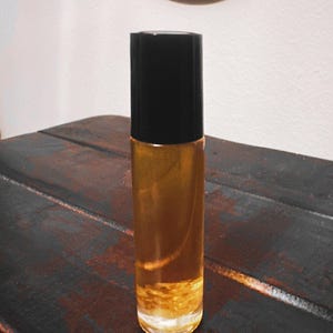 Puede incluir: Un frasco roll-on de vidrio transparente con tapa negra, lleno de líquido dorado. El frasco está sobre una superficie de madera oscura y texturizada. El fondo incluye una pared blanca y un marco circular decorativo.