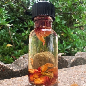 May include: A clear glass bottle with a black lid filled with a light yellow liquid and dried rose petals.