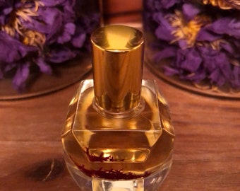 May include: A small, clear glass bottle with a gold cap and a gold label. The bottle is filled with a golden liquid and contains red saffron threads.