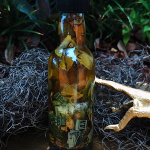Op de afbeelding: Een glazen fles met een zwarte dop gevuld met een gouden vloeistof en verschillende kruiden en dollarbiljetten. De fles staat op een gouden oppervlak met kleine, witte stenen.