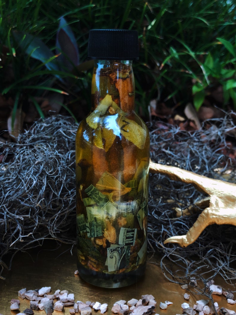 Op de afbeelding: Een glazen fles met een zwarte dop gevuld met een gouden vloeistof en verschillende kruiden en dollarbiljetten. De fles staat op een gouden oppervlak met kleine, witte stenen.