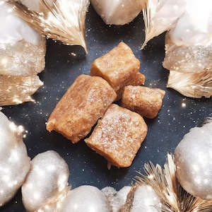 May include: Close-up of four golden brown fudge squares on a black surface. The fudge is dusted with powdered sugar and surrounded by gold and white decorative elements.