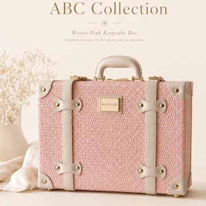 May include: A rectangular pink woven keepsake box with a top handle, featuring cream-colored faux leather straps, gold-tone metal corner reinforcements, and a central gold plaque engraved with the text DAISY FIELDS ABC Collection.