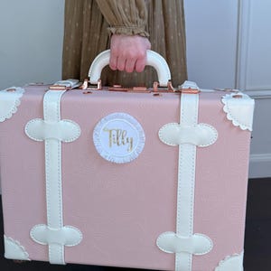 May include: A pink vintage-style suitcase with white trim and a white luggage tag that says "Tilly" in gold lettering.